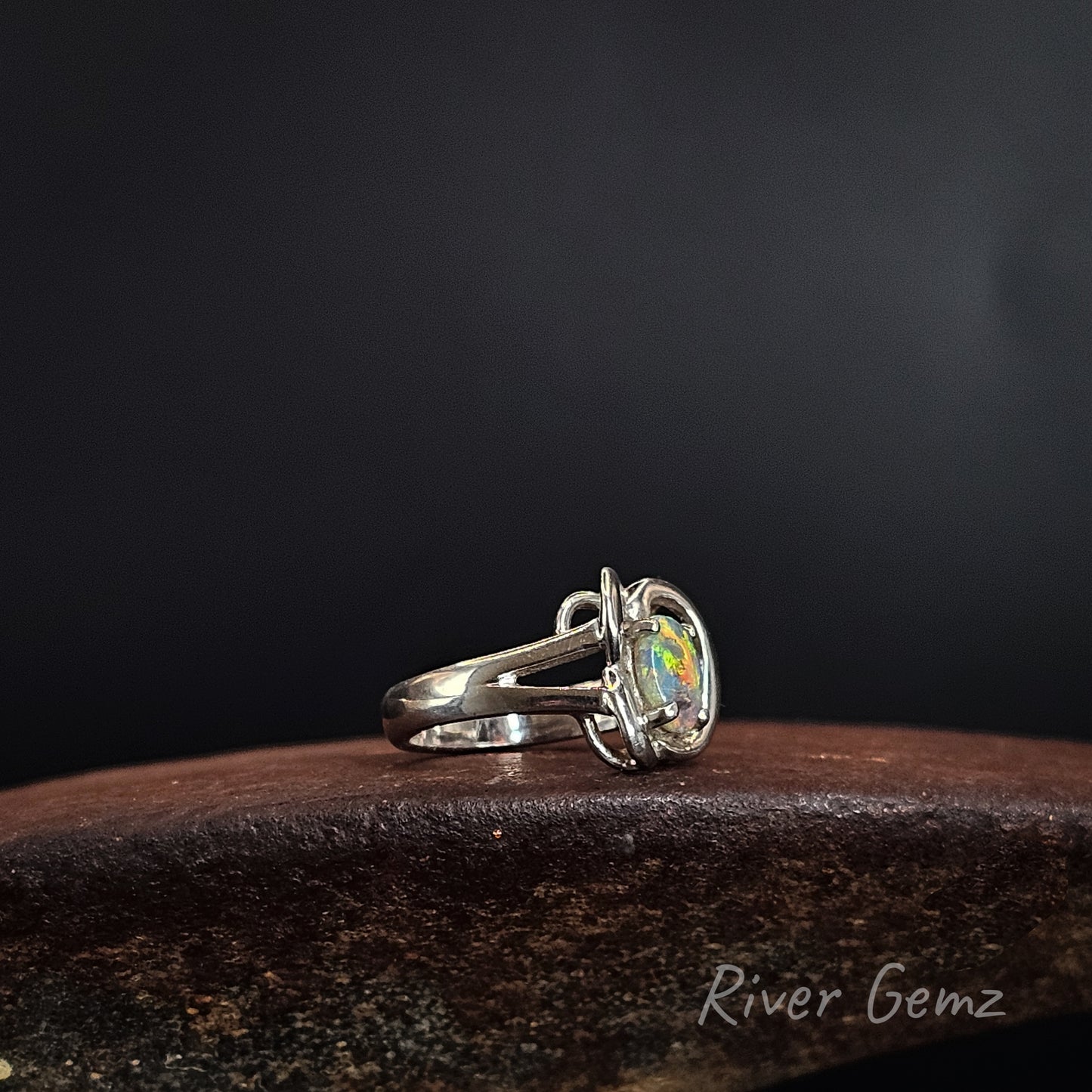 A sterling silver ring with a round iridescent opal gemstone, resting on a dark textured wooden surface against a dark gray background. The brand name 'River Gemz' is visible in the bottom right corner.