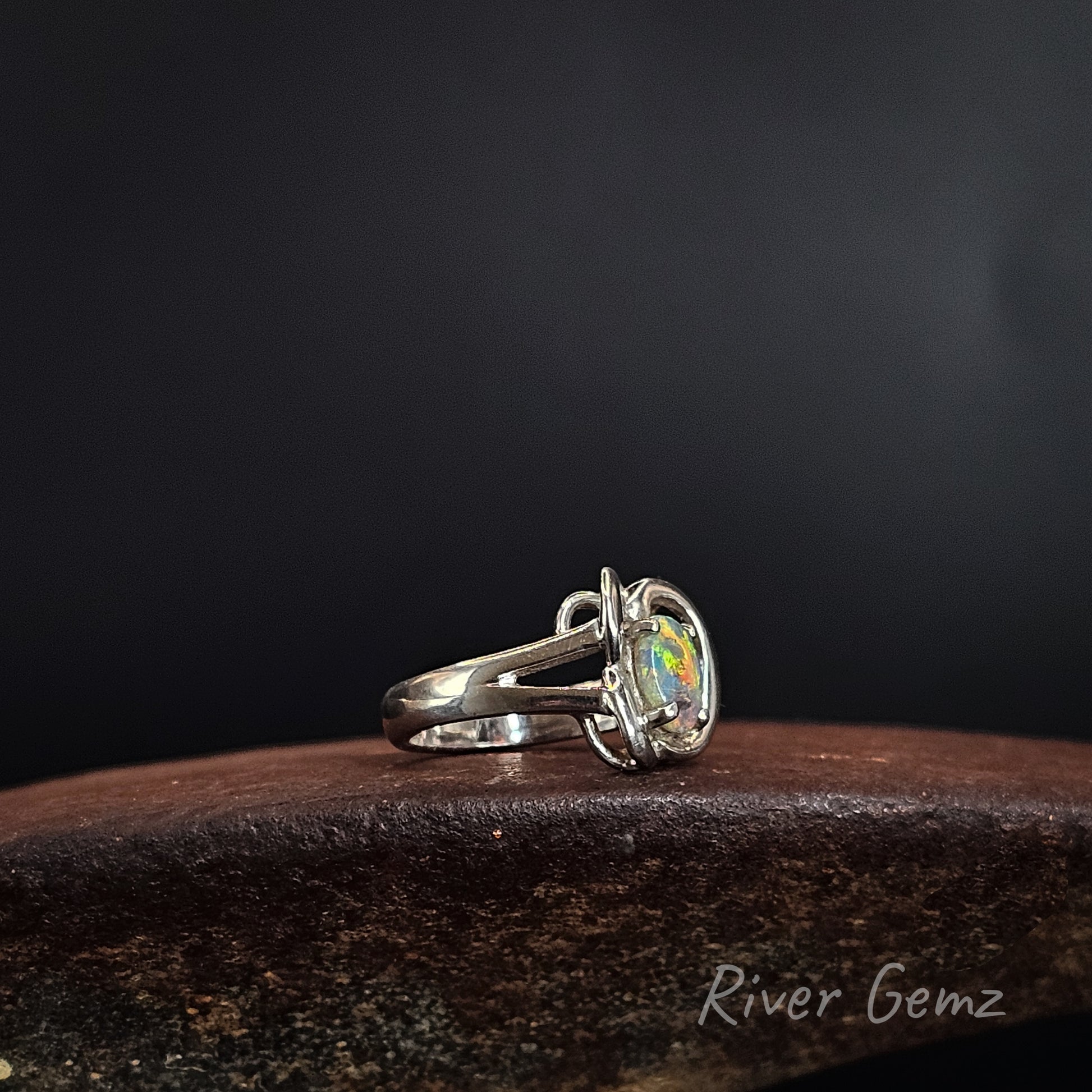 A sterling silver ring with a round iridescent opal gemstone, resting on a dark textured wooden surface against a dark gray background. The brand name 'River Gemz' is visible in the bottom right corner.