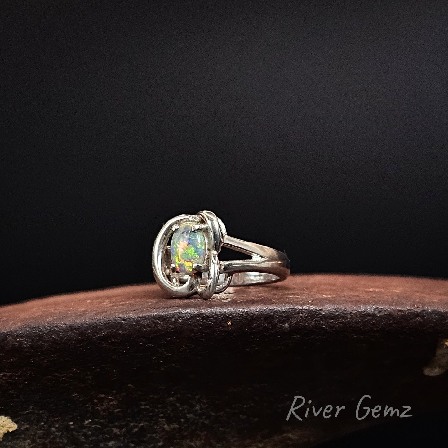 A sterling silver ring featuring a round iridescent opal gemstone, resting on a dark brown textured surface against a black background. The brand name 'River Gemz' is visible in the bottom right corner.