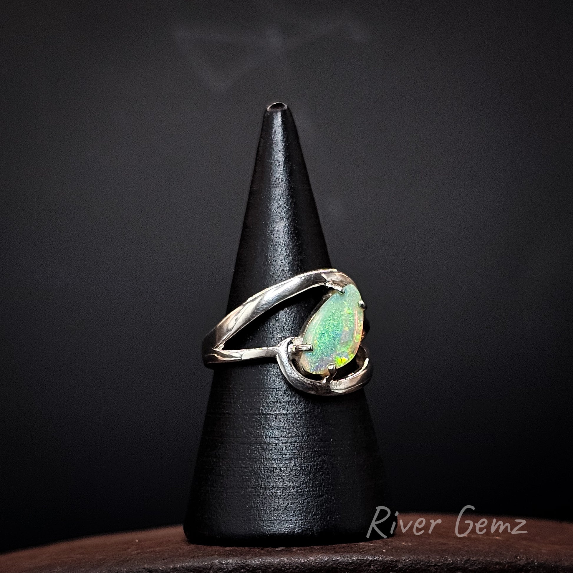 A sterling silver ring with a teardrop-shaped iridescent opal gemstone, displayed on a black cone stand against a dark background. The brand name 'River Gemz' is visible in the bottom right corner.