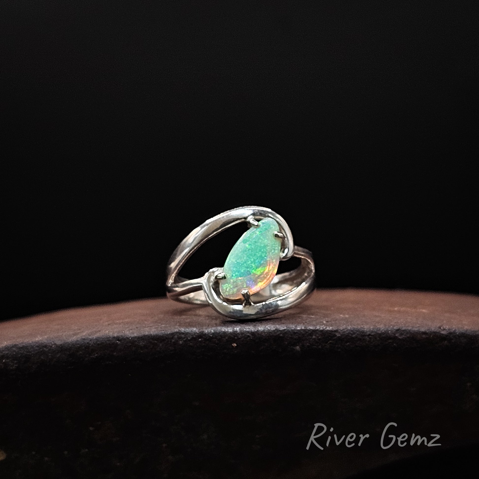 A sterling silver ring featuring a claw-set opal gemstone with vibrant green, orange, and blue flashes, displayed on a dark wooden surface against a black background. The brand name 'River Gemz' is visible in the bottom right corner.