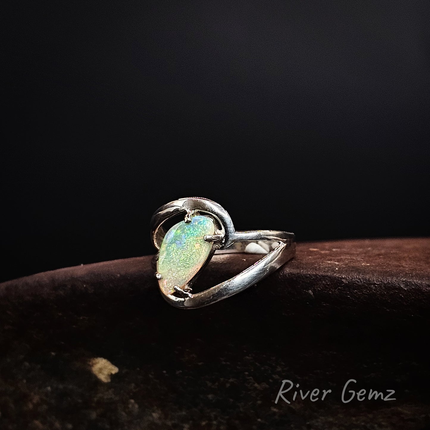 A sterling silver ring featuring a triangular-shaped iridescent opal gemstone, resting on a dark brown textured surface against a black background. The brand name 'River Gemz' is visible in the bottom right corner.