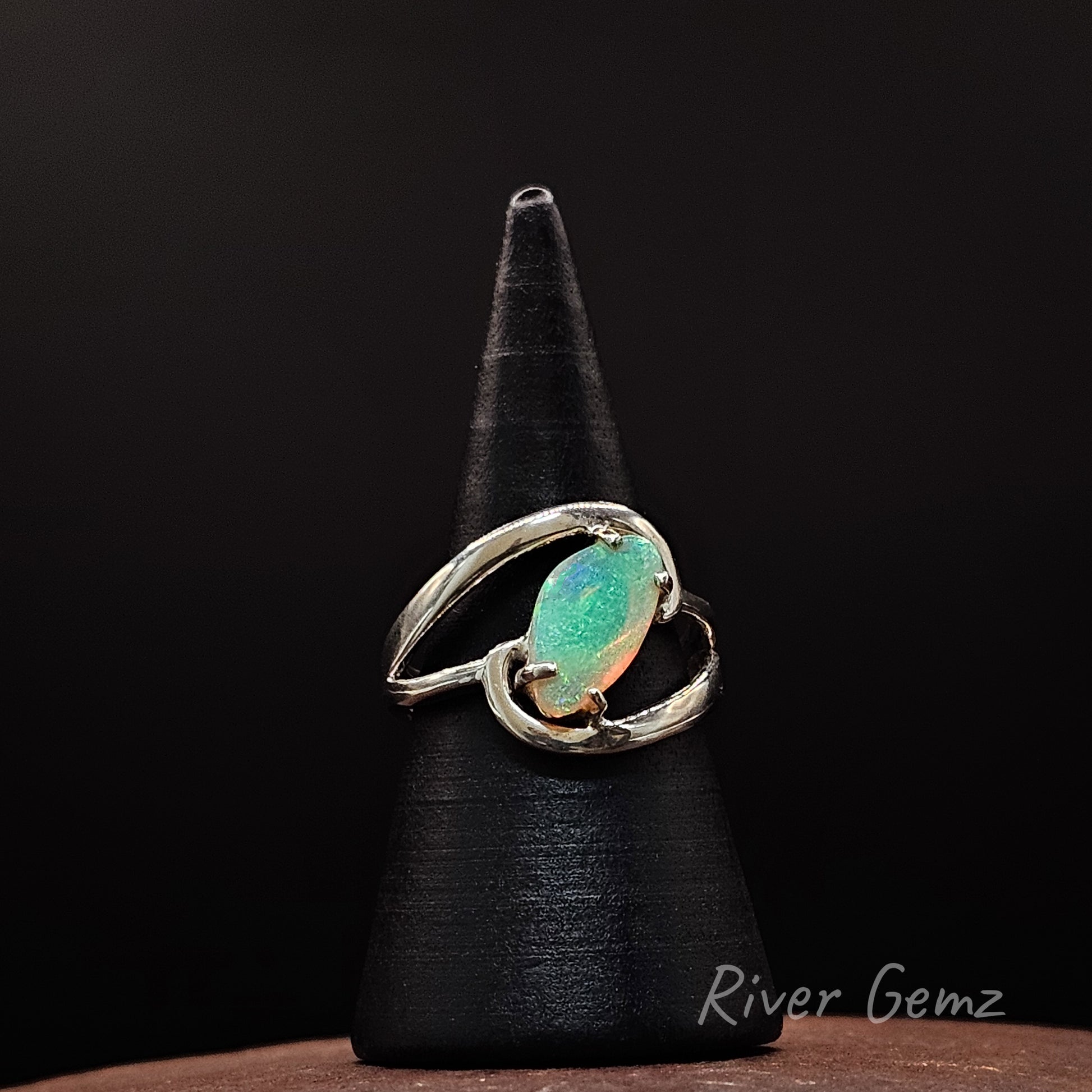 A silver ring with a free-form cut iridescent green opal gemstone, displayed on a black cone stand against a dark background. The brand name 'River Gemz' is visible in the bottom right corner.