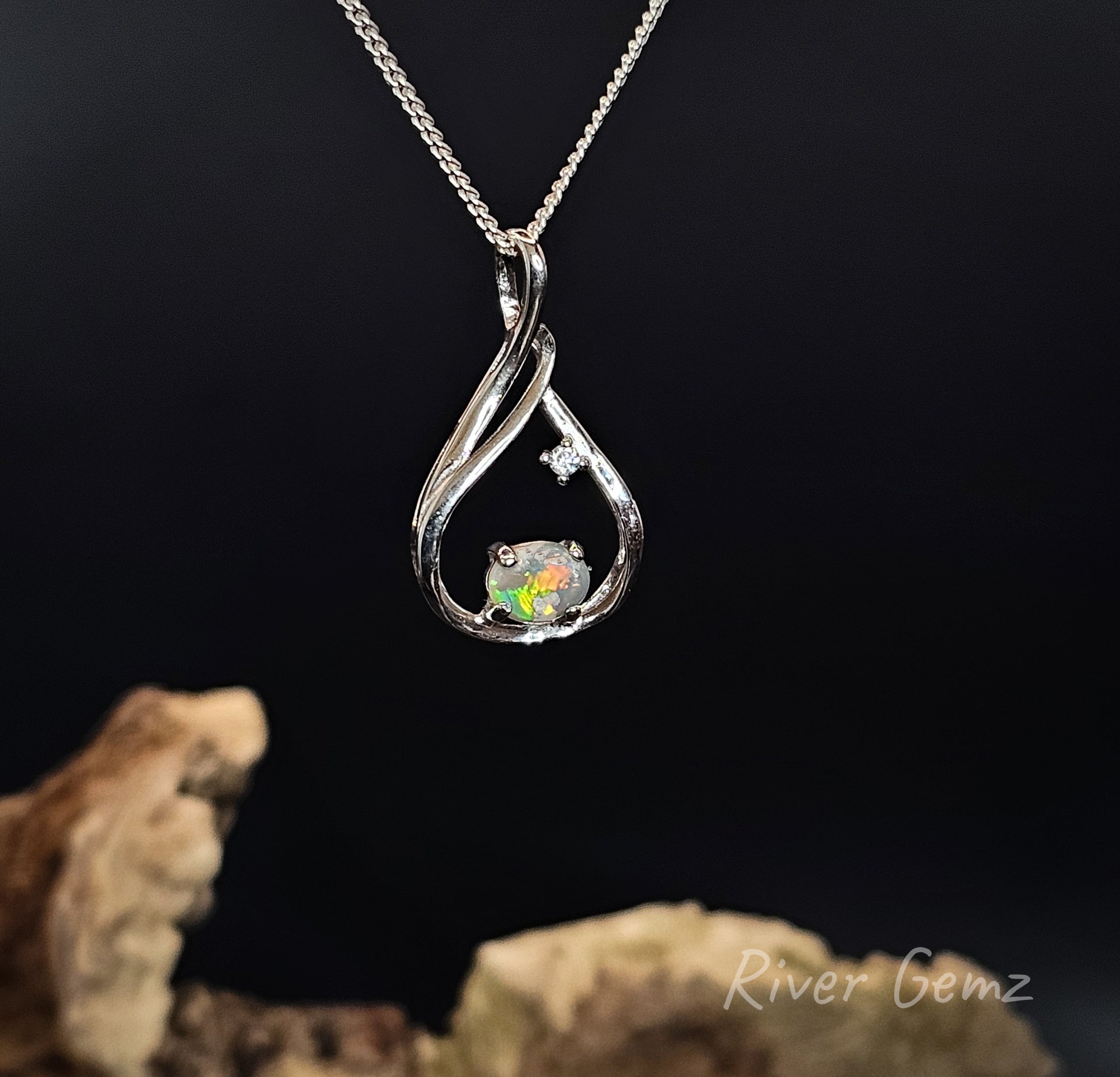 A silver teardrop-shaped pendant necklace featuring a small colorful iridescent opal and a tiny clear gemstone, displayed against a dark background with driftwood props and a River Gemz watermark.