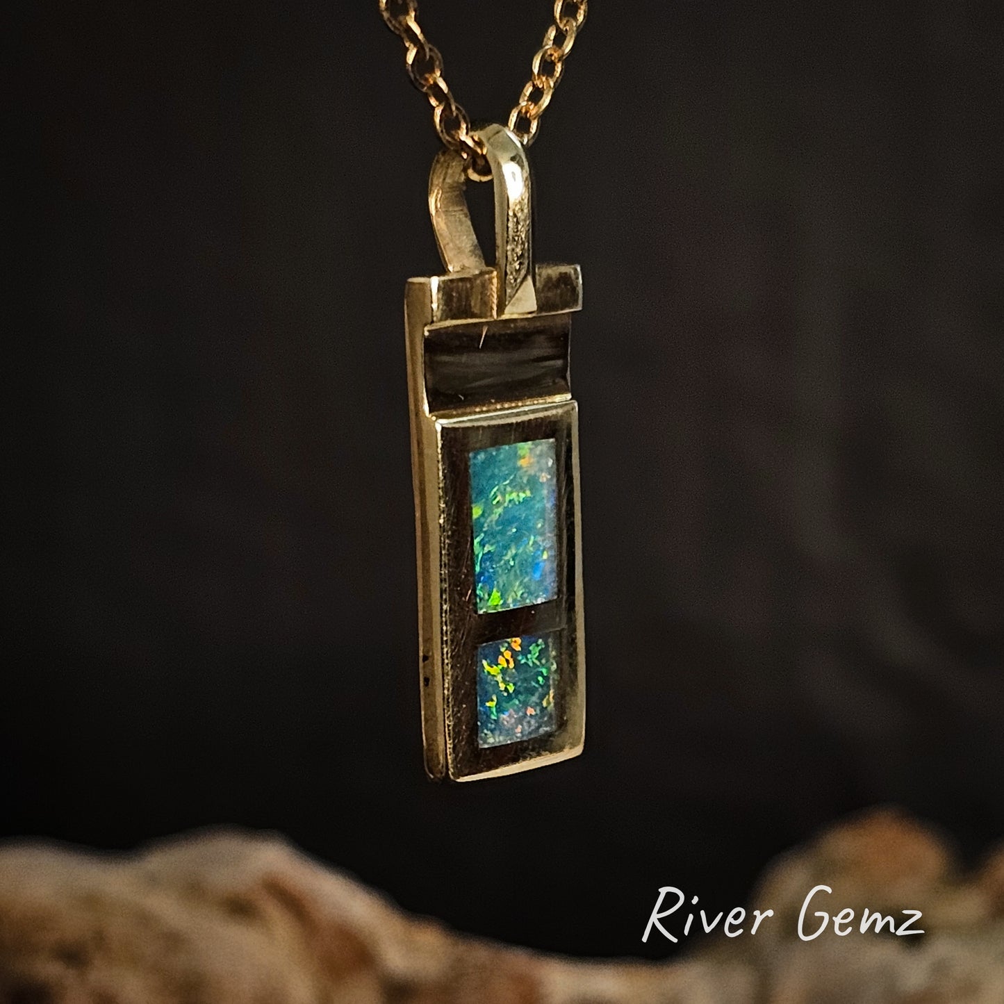 Gold pendant with a colourful gemstone on a dark background, featuring 'River Gemz' branding.