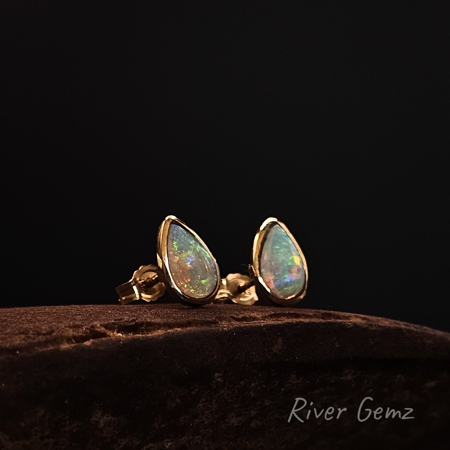 Crystal opals in gold stud earrings on a dark surface with 'River Gemz' branding.