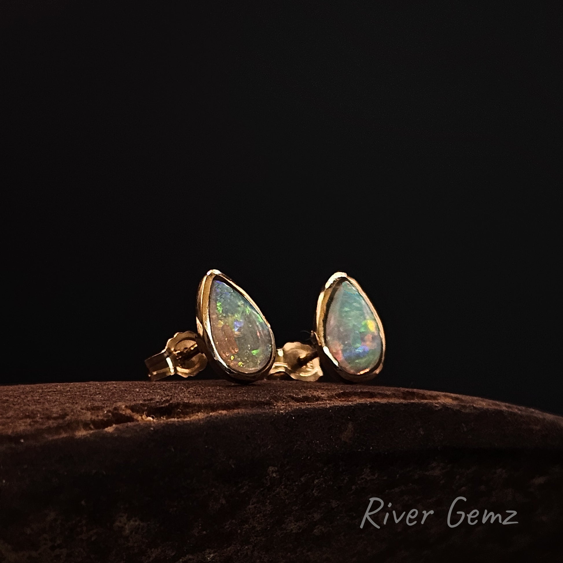 Crystal opals in gold stud earrings on a dark surface with 'River Gemz' branding.