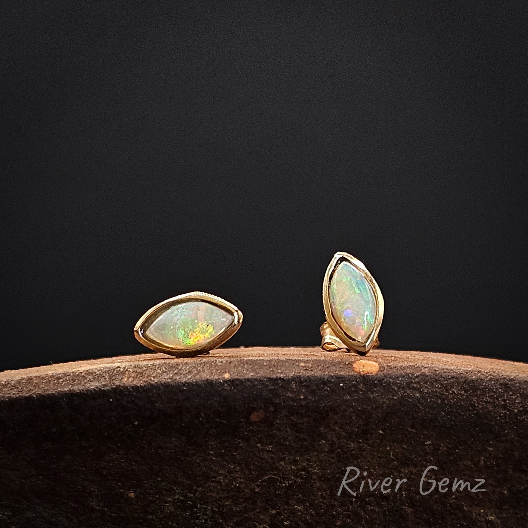 Light opals besel set in yellow gold earrings on a dark surface with 'River Gemz' branding.