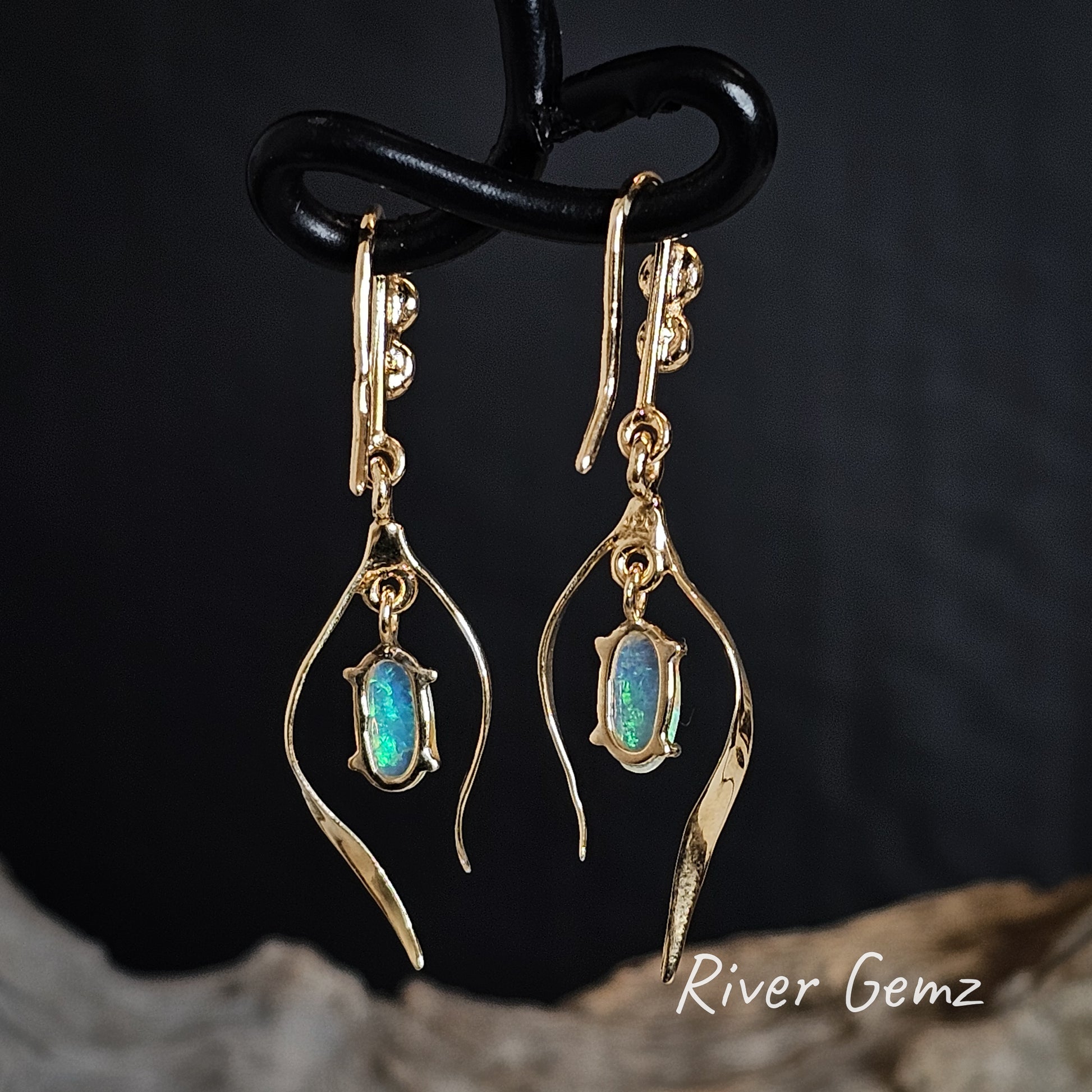 Back of gold earrings with crystal opal stones on a dark background, featuring the brand 'River Gemz'.
