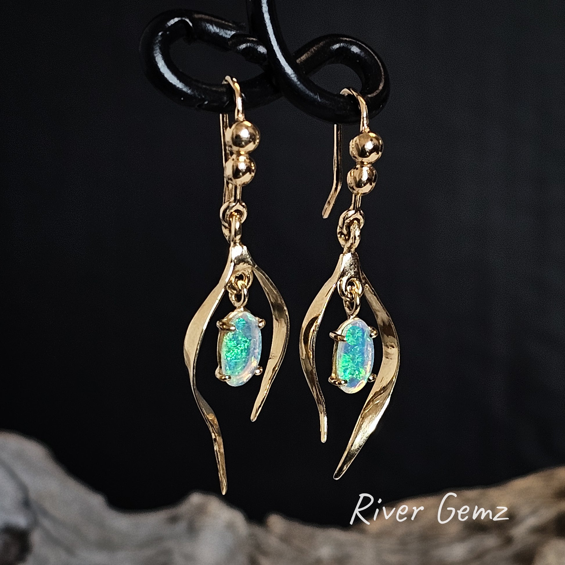 Gold shepherd hook earrings with green crystal opals on a dark background, featuring the brand 'River Gemz'.