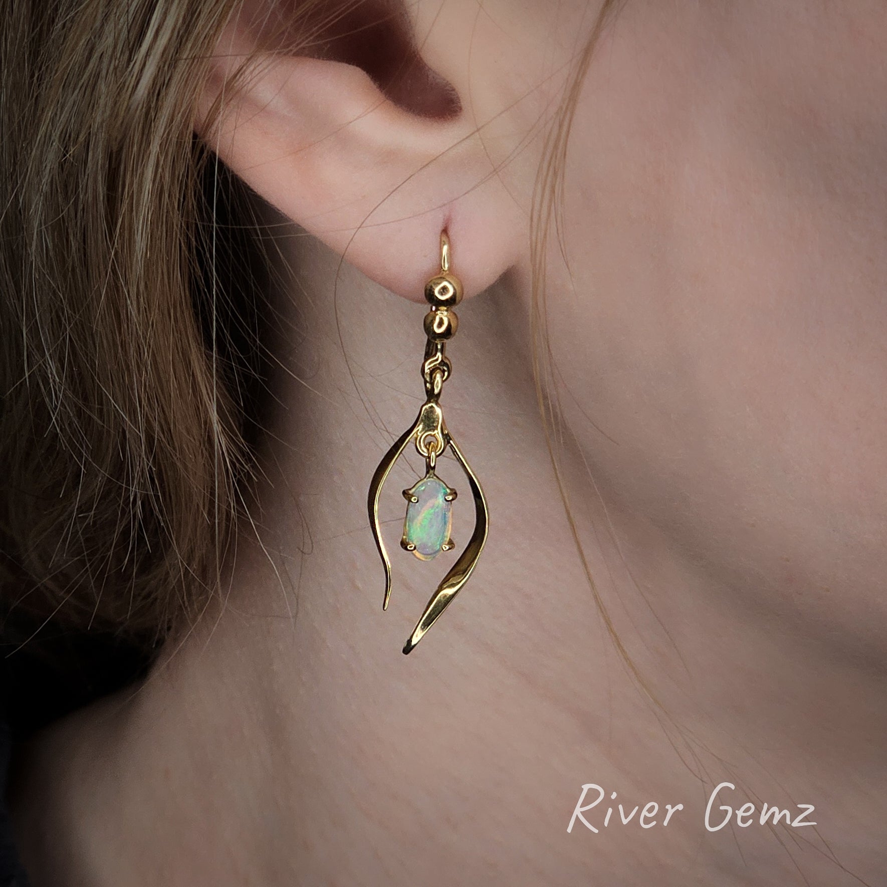 Yellow gold dangly earring with a crystal opal worn by a person, close-up of ear.