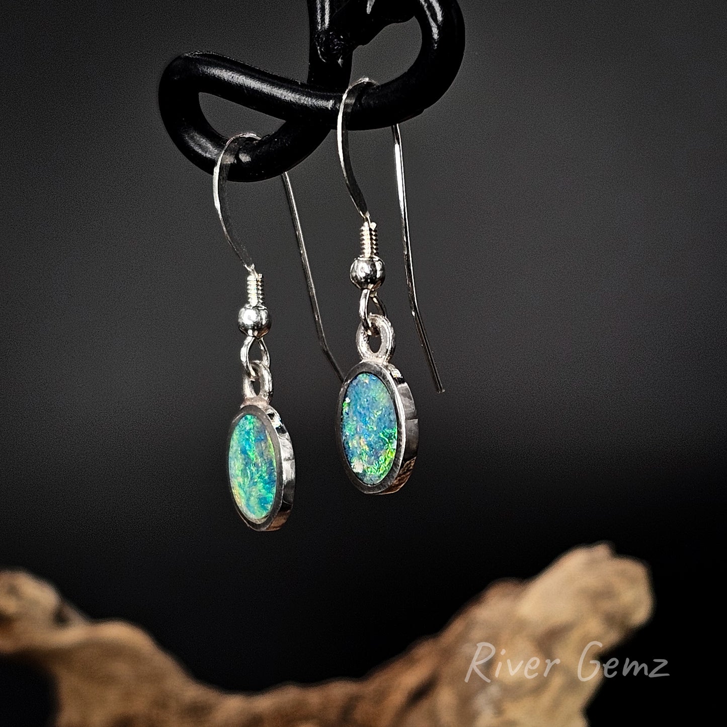 Opal earrings hanging on a black hook with a dark background and 'River Gemz' branding.