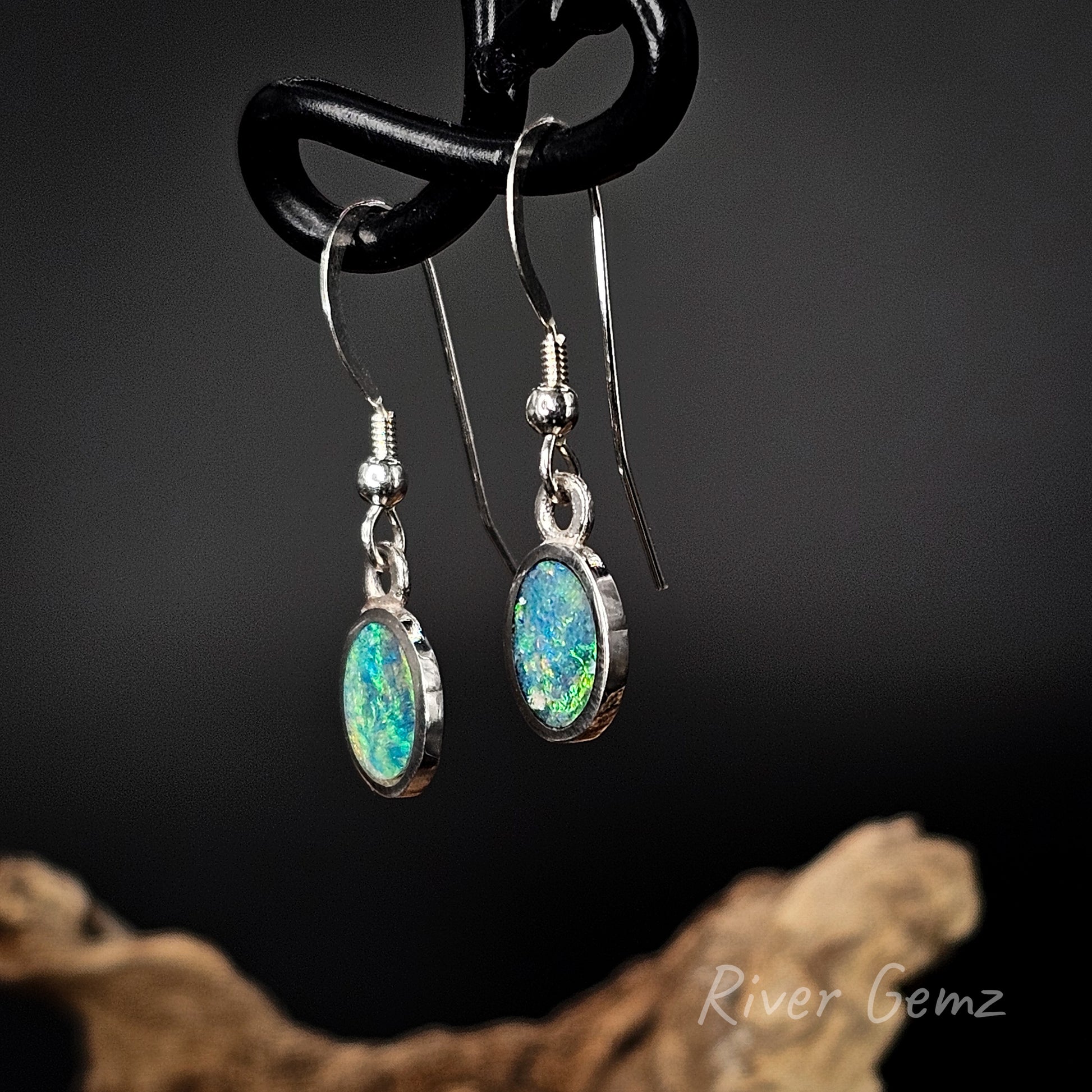 Opal earrings hanging on a black hook with a dark background and 'River Gemz' branding.