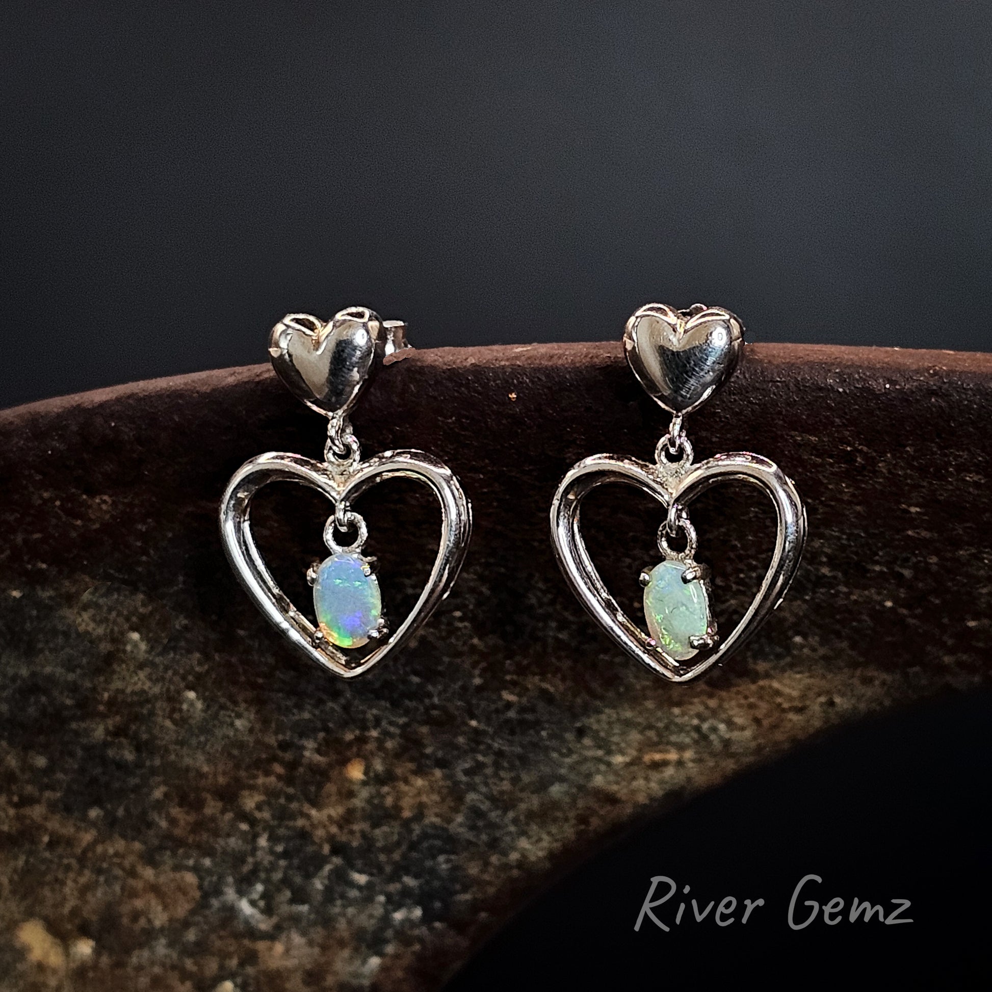 Heart-shaped earrings with opal stones on a dark background, branded 'River Gemz'.