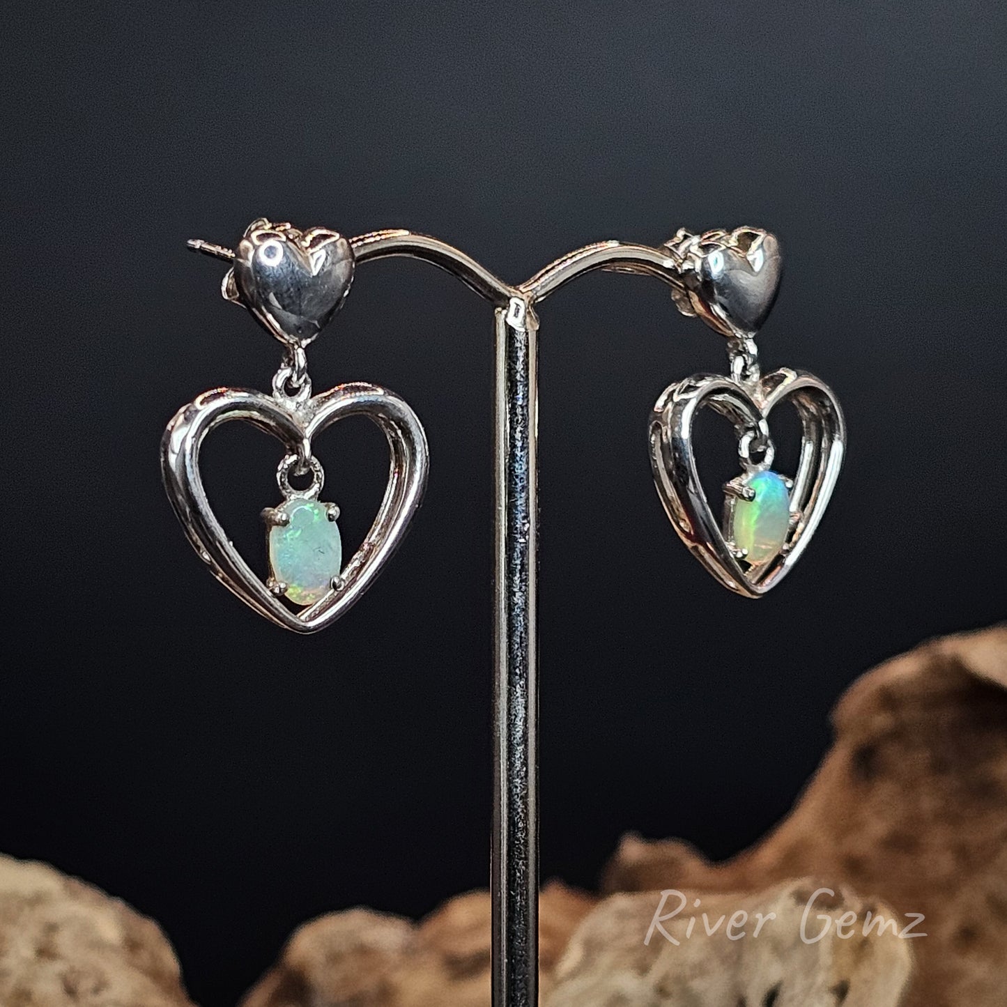 Heart-shaped earrings with opal stones on a stand against a dark background.