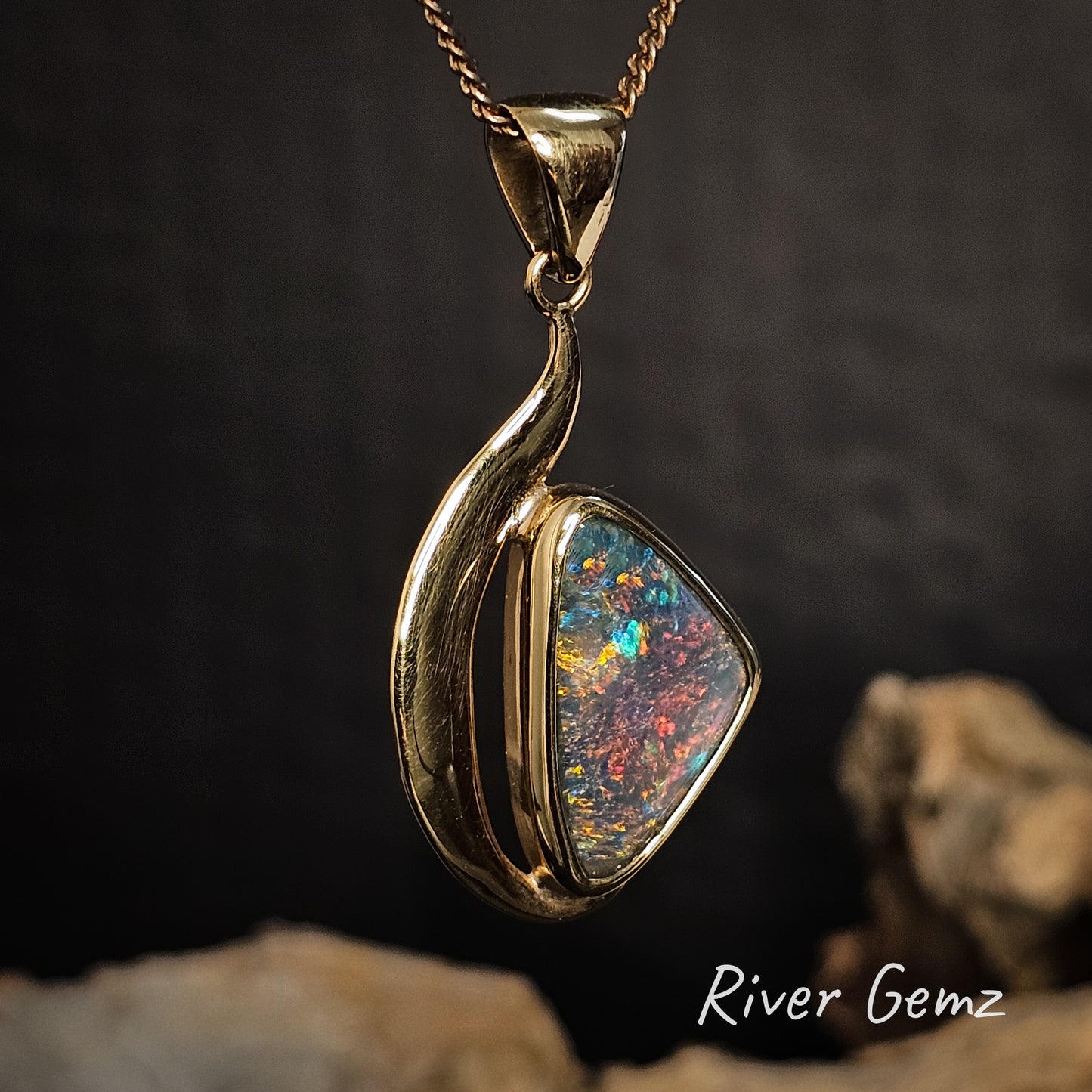 Gold pendant with a triangular shaped semi-black opal on a dark background, featuring 'River Gemz' branding.