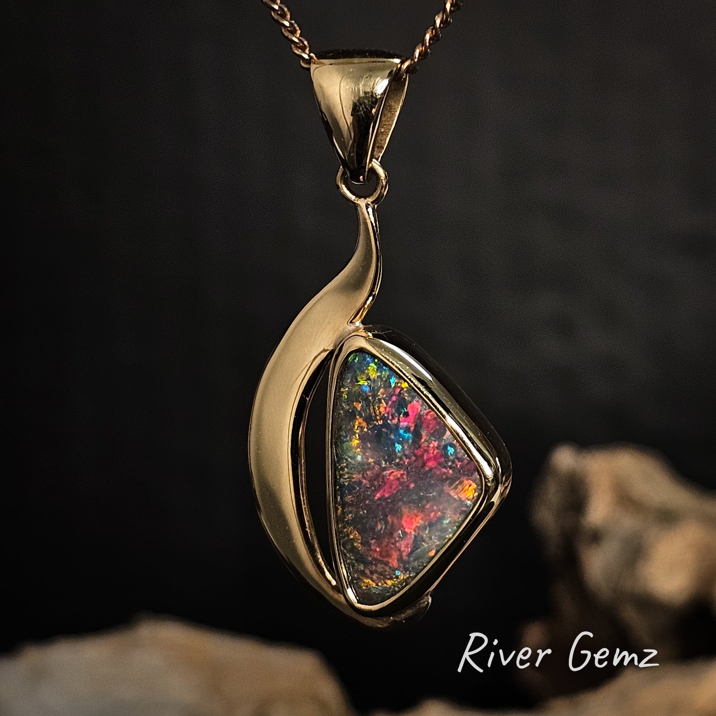 Gold pendant with a colorful gemstone on a dark background, featuring 'River Gemz' branding.