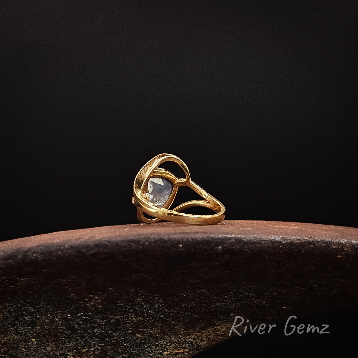 Rear view of gold ring showing the dark base tone of the semi-black opal set in it on a weathered shoe anvil, black background and 'River Gemz' brand in the bottom corner.