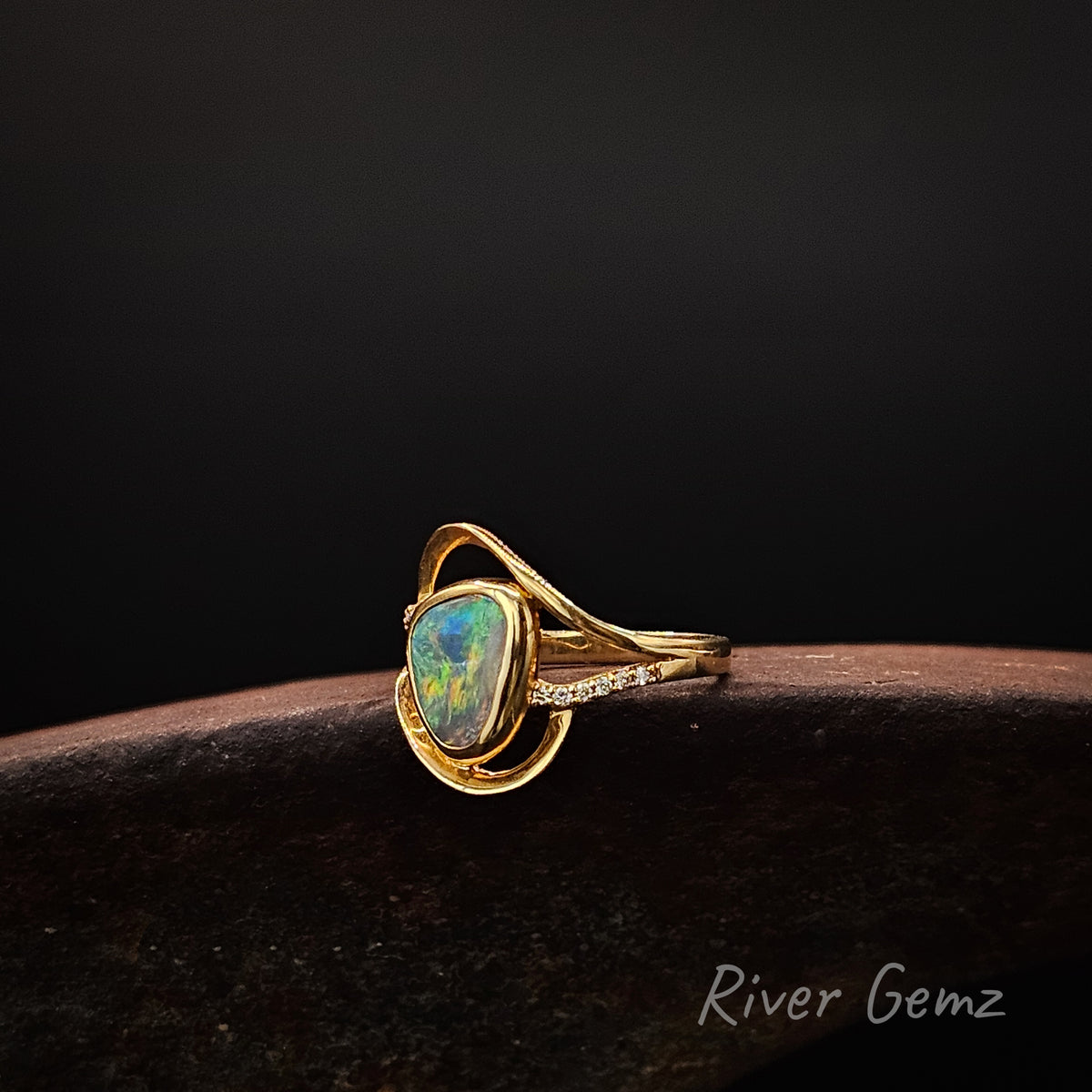 Yellow gold ring with central opal and white diamonds on the shoulders on a weathered shoe anvil, black background and 'River Gemz' brand in the bottom corner.