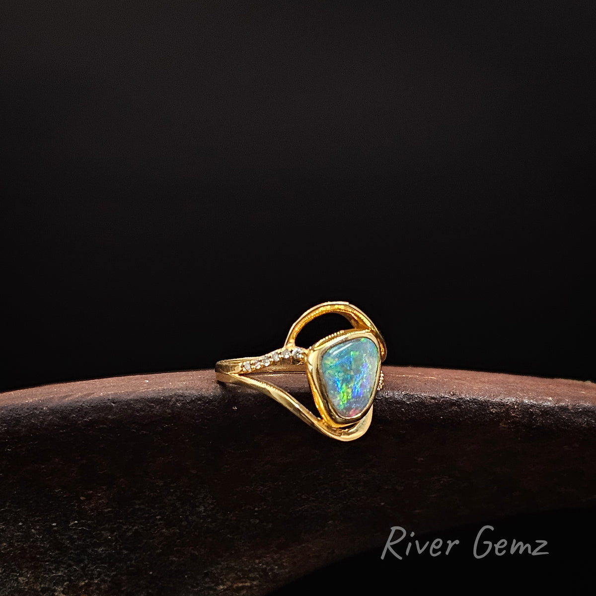 Yellow gold ring with central opal and white diamonds on the shoulders on a weathered shoe anvil, black background and 'River Gemz' brand in the bottom corner.
