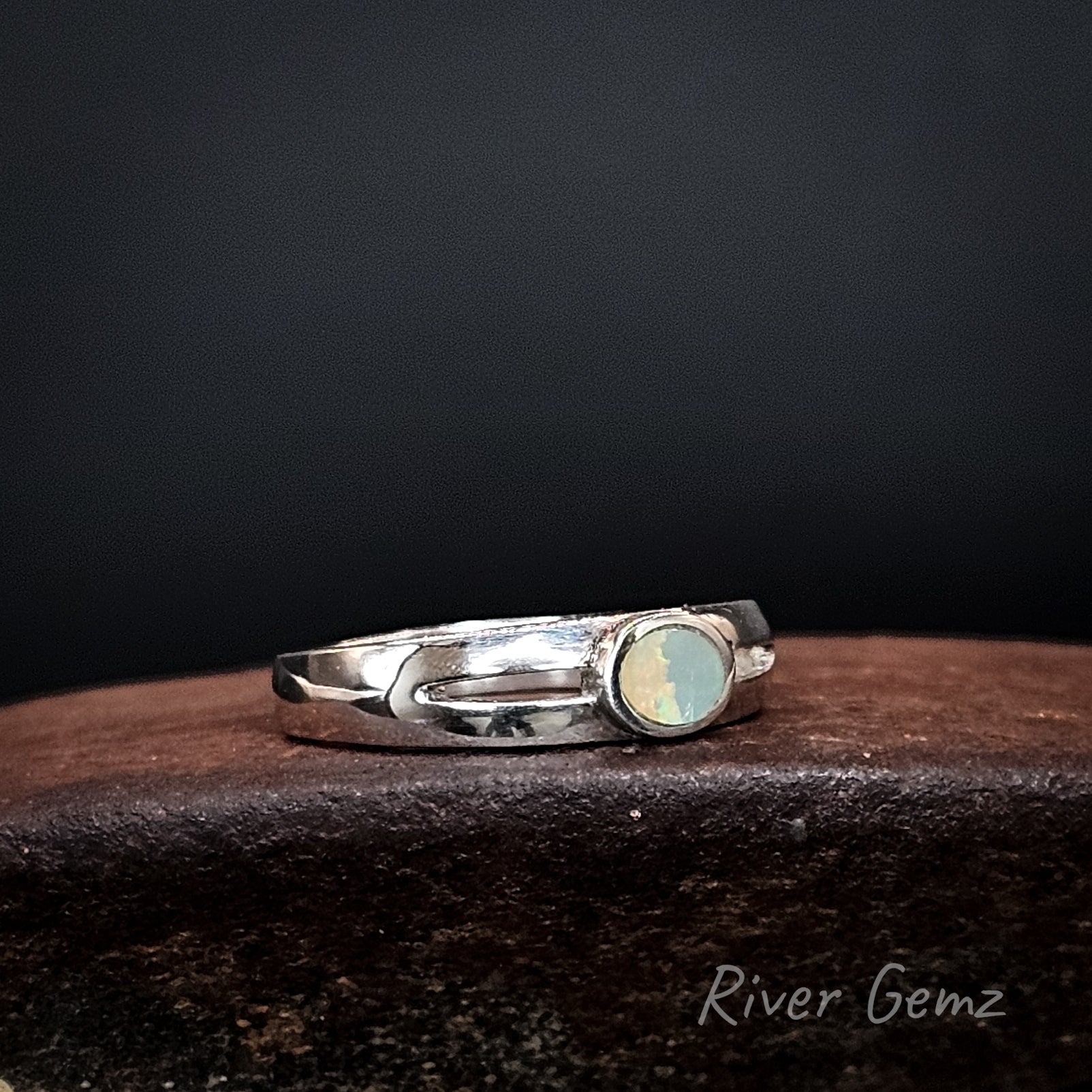 Silver ring with an oval shaped dark opal on a brown coloured base with 'River Gemz' branding.
