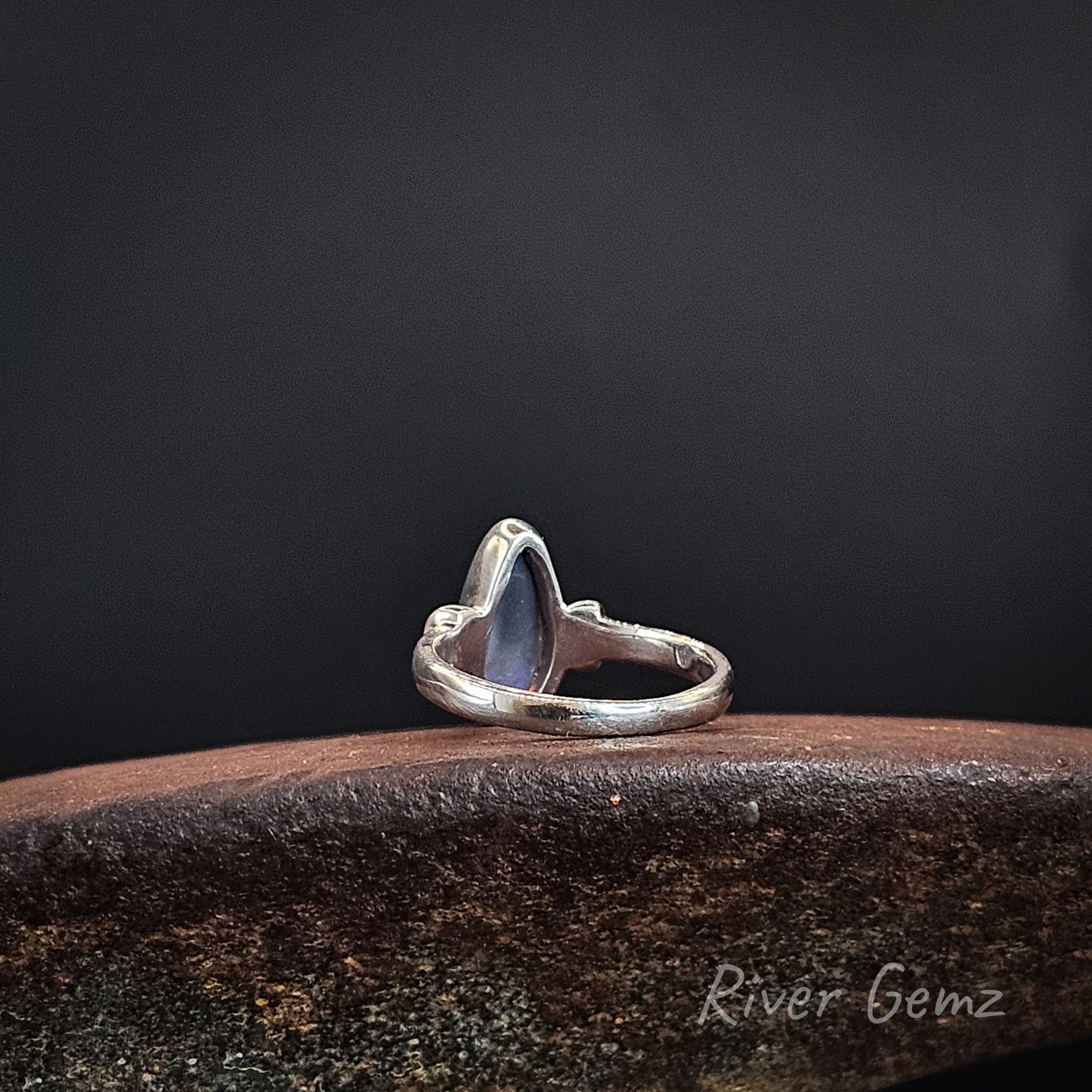 Rear view of the ring shows the dark base tone of the semi-black opal, resting on a textured dark brown wooden surface against a dark gray background. The brand name 'River Gemz' is visible as a watermark in the bottom right corner.