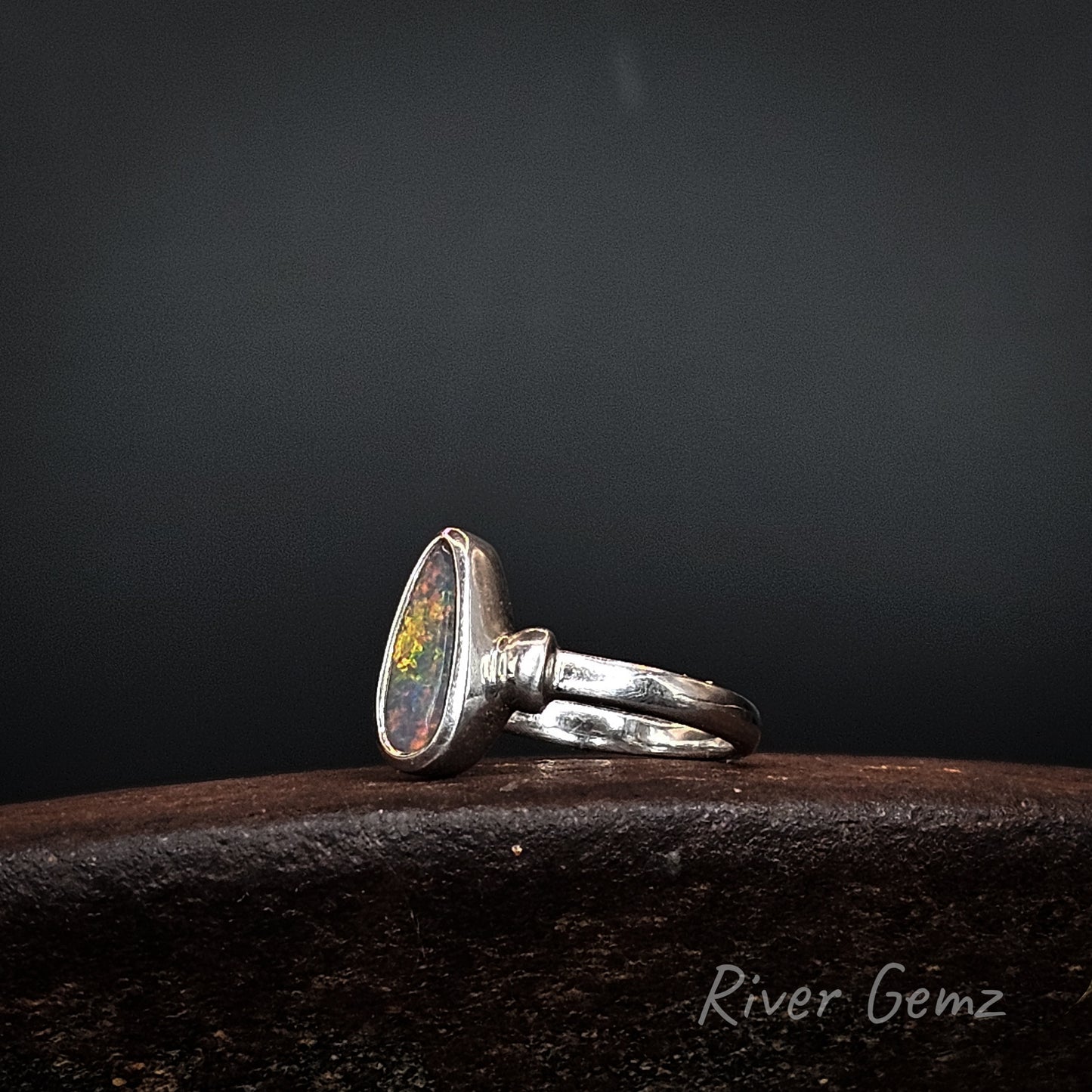 A sterling silver ring featuring a teardrop-shaped cabochon opal gemstone with vibrant multicolor play-of-color, resting on a dark textured wooden surface against a dark gray background.