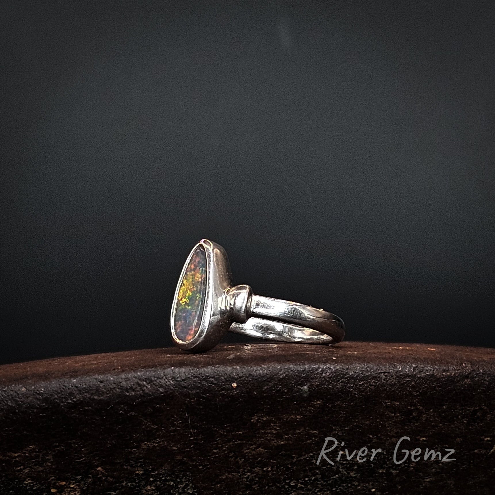A sterling silver ring featuring a teardrop-shaped cabochon opal gemstone with vibrant multicolor play-of-color, resting on a dark textured wooden surface against a dark gray background.