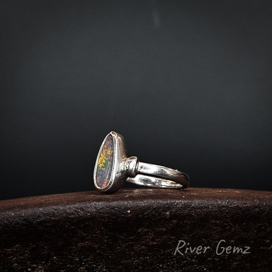 A sterling silver ring featuring a teardrop-shaped cabochon opal gemstone with vibrant multicolor play-of-color, resting on a dark textured wooden surface against a dark gray background.