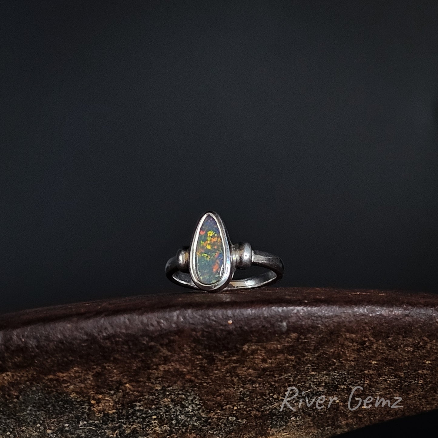 A sterling silver ring featuring a teardrop-shaped opal gemstone with vibrant multicolor play-of-color, displayed on a dark wooden surface against a dark background.