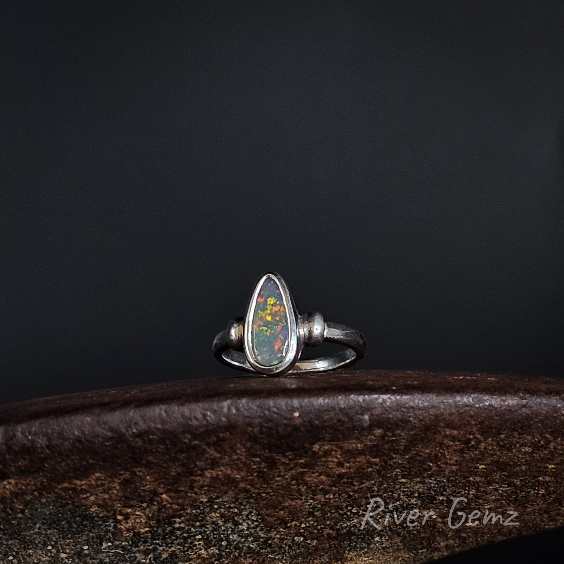 A sterling silver ring featuring a teardrop-shaped opal gemstone with vibrant multicolor play-of-color, displayed on a dark wooden surface against a dark background.