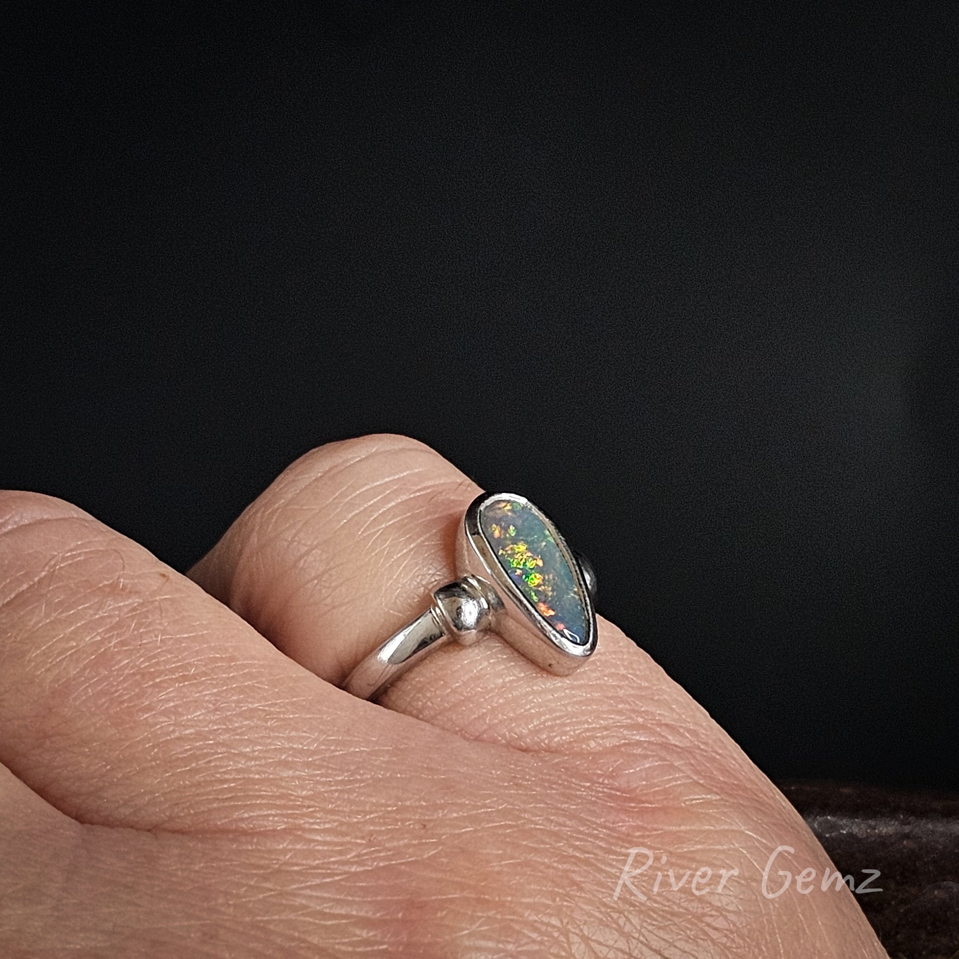 A silver ring with an oval opal gemstone featuring a play-of-color pattern, worn on a person's finger against a dark background.