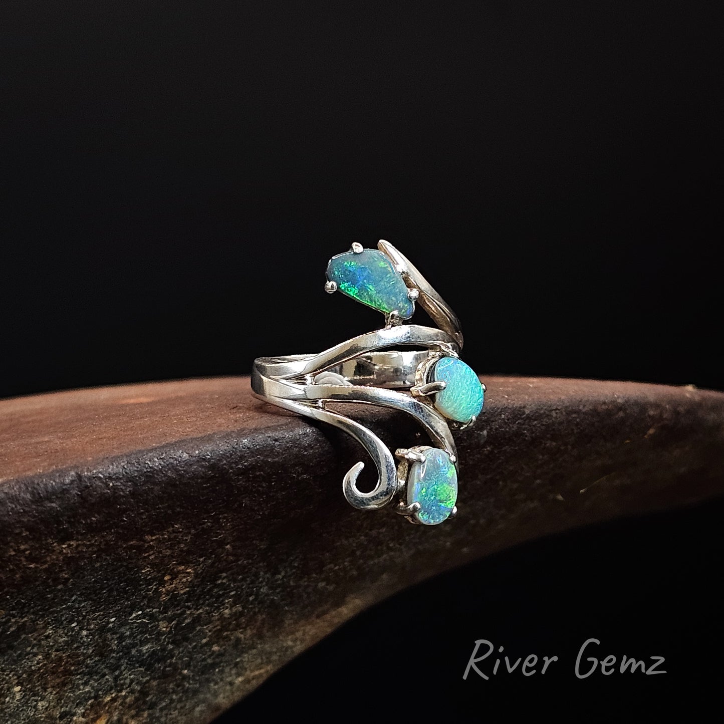 A sterling silver ring with three iridescent opal gemstones in a flowing, wave-like setting, displayed on a dark wooden surface against a black background. The brand name 'River Gemz' is visible in the bottom right corner.
