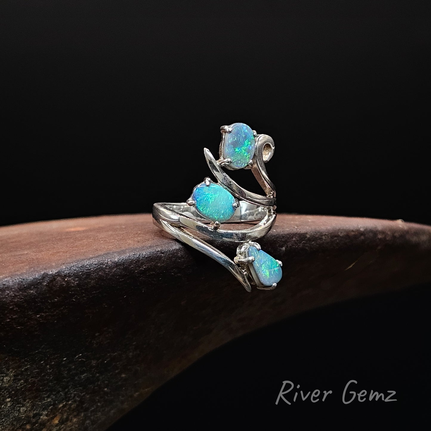 A silver ring featuring three blue opal gemstones, displayed on a dark wooden surface against a black background. The brand name 'River Gemz' is visible in the bottom right corner.
