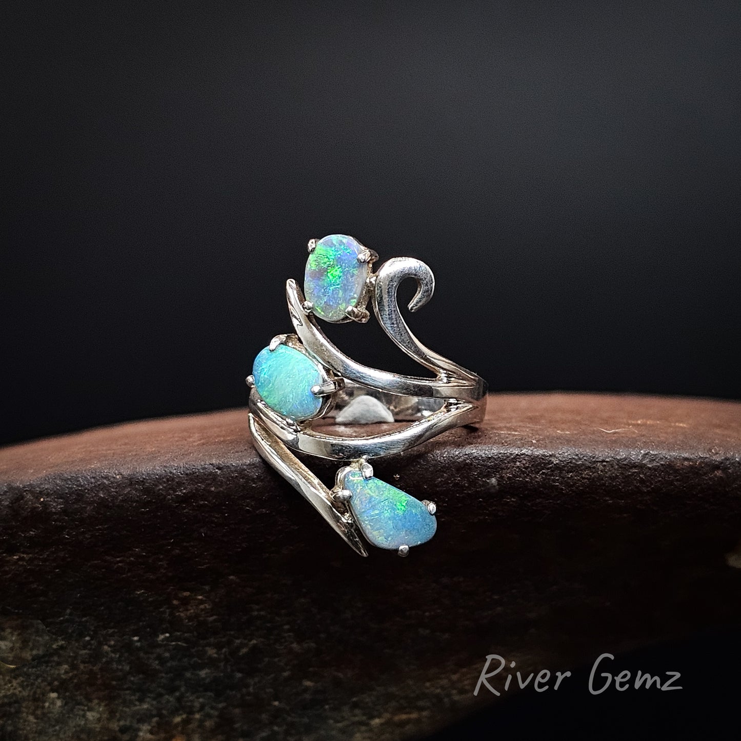 A silver-toned wrap ring featuring three teardrop-shaped iridescent blue-green opal gemstones, displayed on a dark wooden surface against a black background. The brand name 'River Gemz' is visible in the bottom right corner.