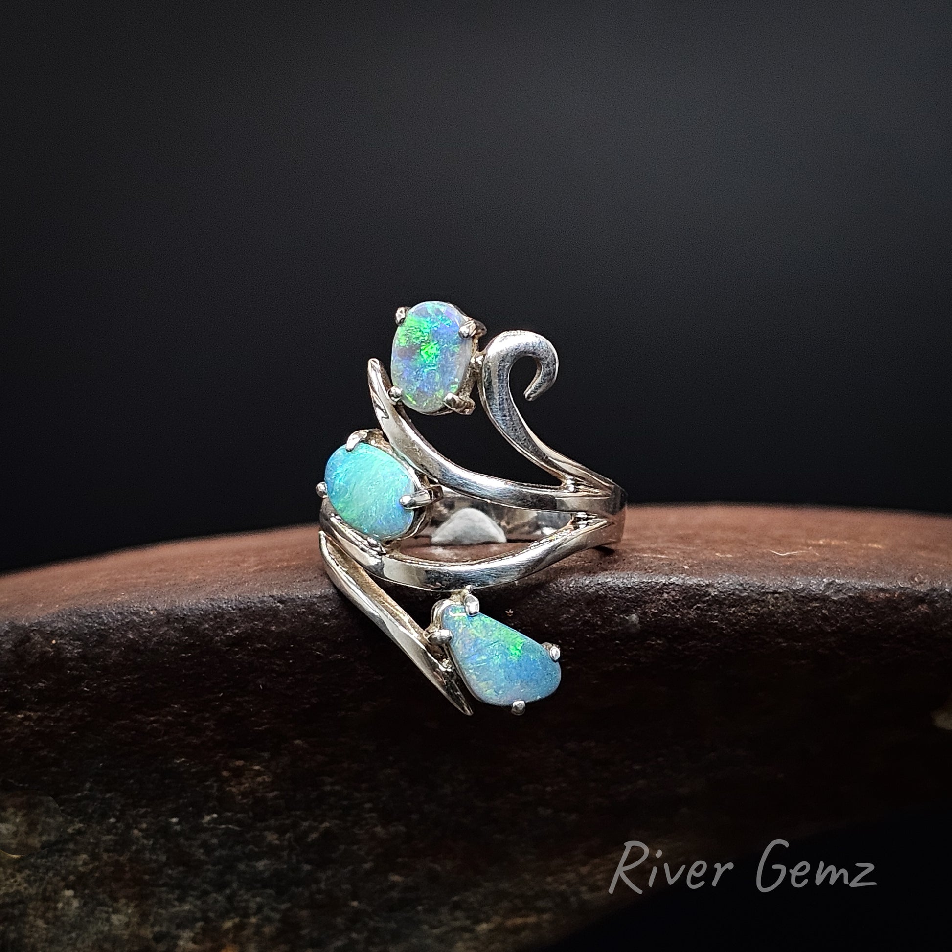 A silver-toned wrap ring featuring three teardrop-shaped iridescent blue-green opal gemstones, displayed on a dark wooden surface against a black background. The brand name 'River Gemz' is visible in the bottom right corner.