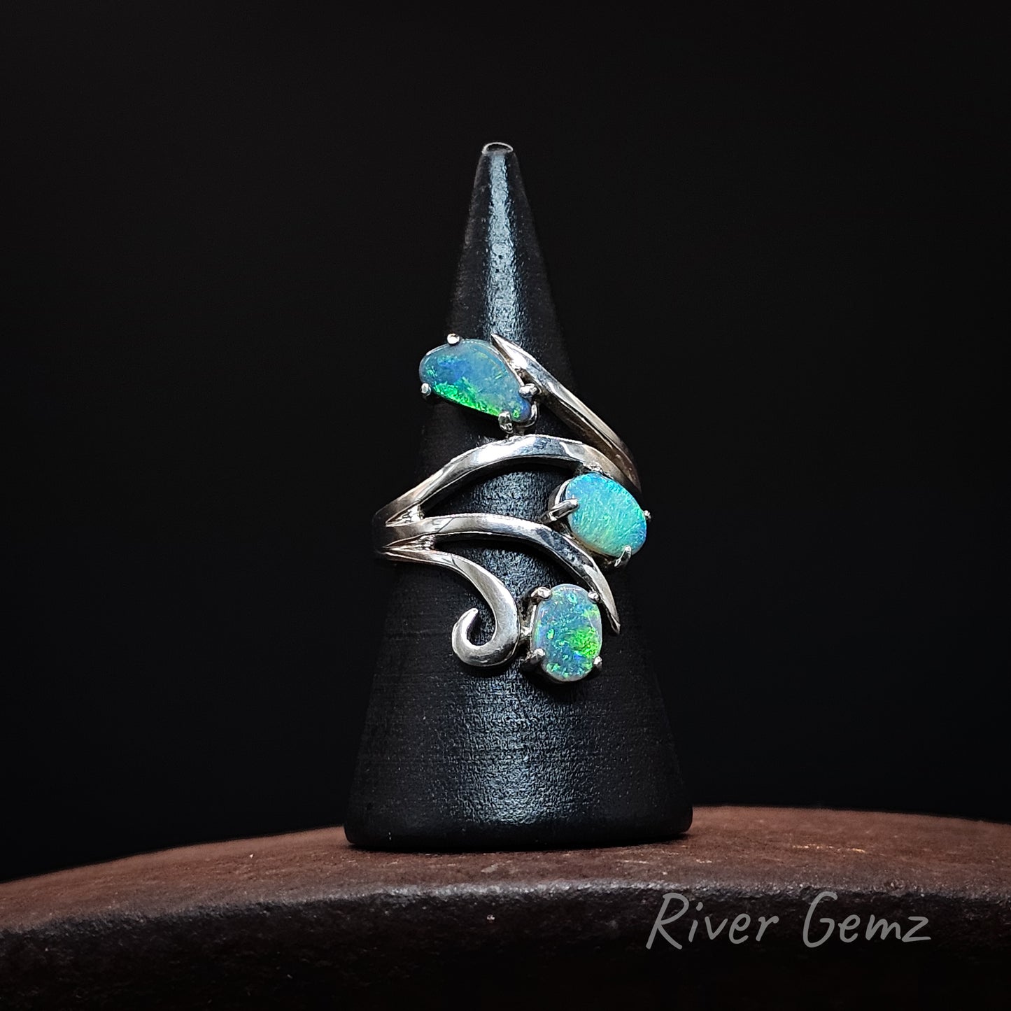 A silver ring with three iridescent blue-green opal gemstones, displayed on a black cone stand over a dark wooden surface. The brand name 'River Gemz' is visible in the bottom right corner.
