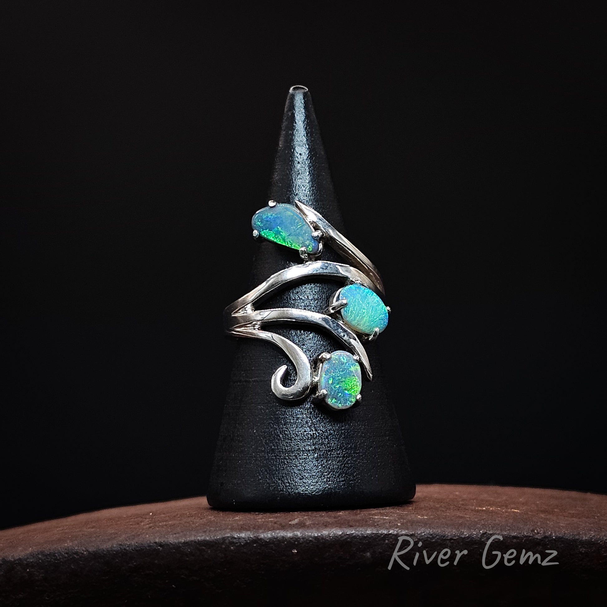 A silver ring with three iridescent blue-green opal gemstones, displayed on a black cone stand over a dark wooden surface. The brand name 'River Gemz' is visible in the bottom right corner.