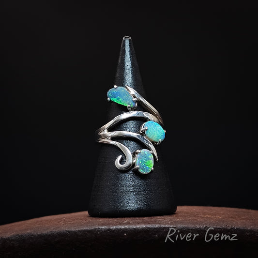 A silver ring with three iridescent blue-green opal gemstones, displayed on a black cone stand over a dark wooden surface. The brand name 'River Gemz' is visible in the bottom right corner.
