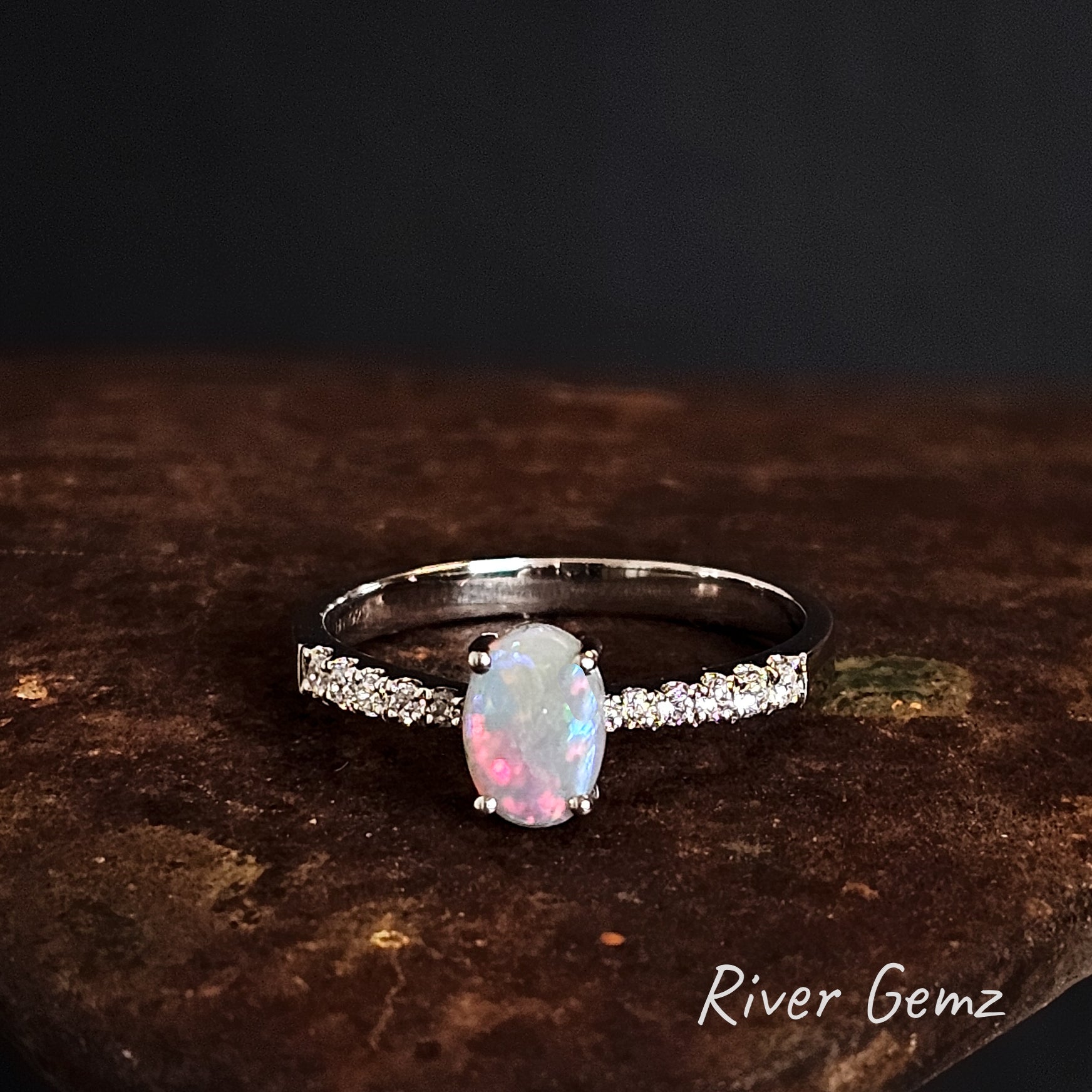 Opal ring with white topaz shoulders on a dark surface, branded 'River Gemz'.