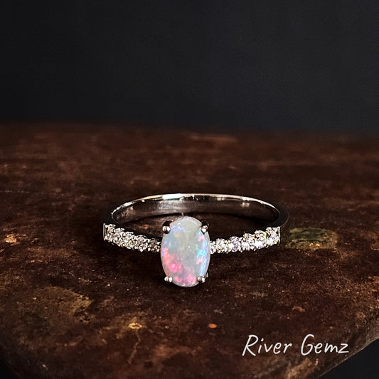 Opal ring with white topaz shoulders on a dark surface, branded 'River Gemz'.