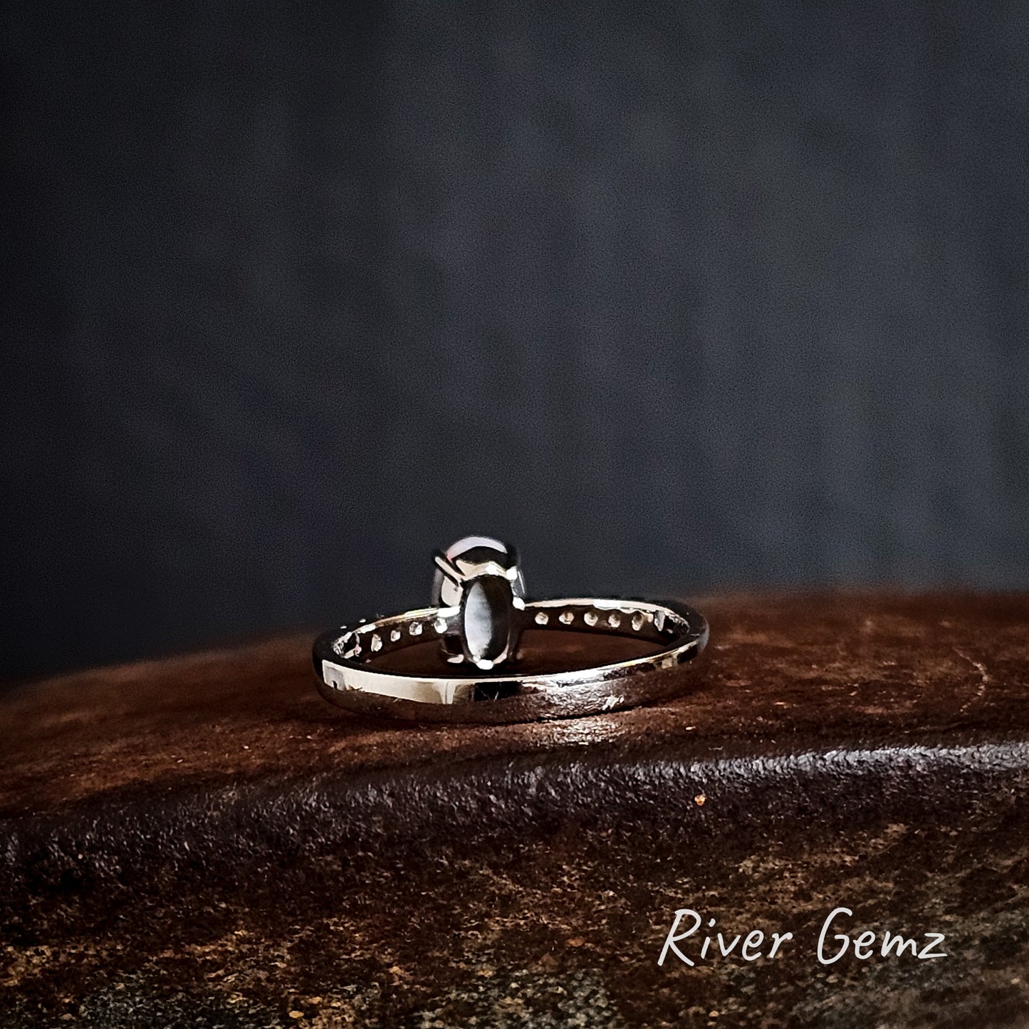 Back view of the silver ring shows the grey tone of the semi-black opal on a rusty brown surface, featuring 'River Gemz' branding.
