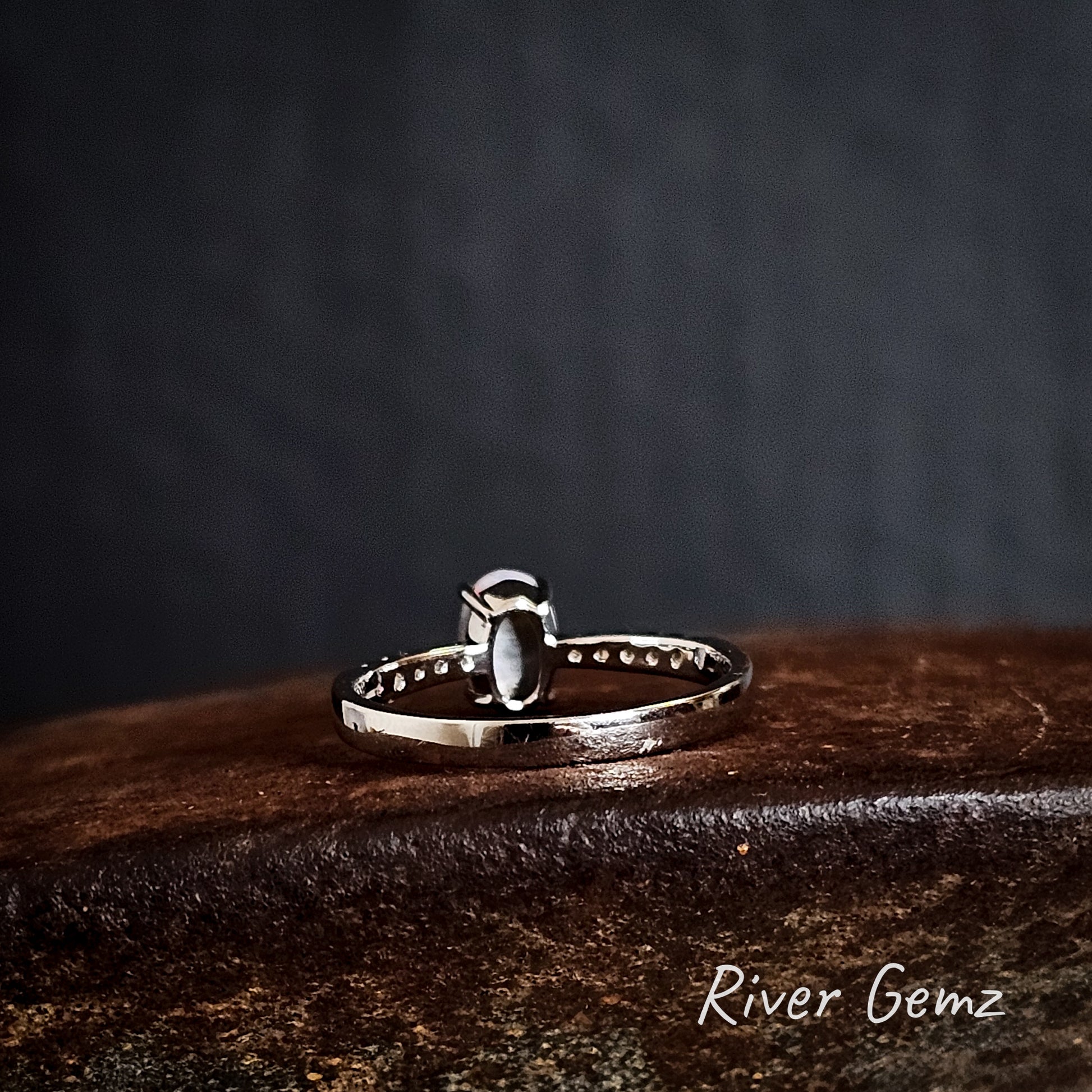 Back view of the silver ring shows the grey tone of the semi-black opal on a rusty brown surface, featuring 'River Gemz' branding.