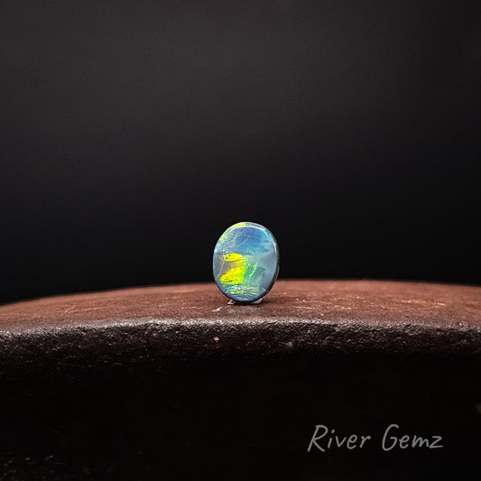 Oval shaped multi-coloured loose opal standing upright on brown surface and black background with 'River Gemz' watermark in bottom right corner.