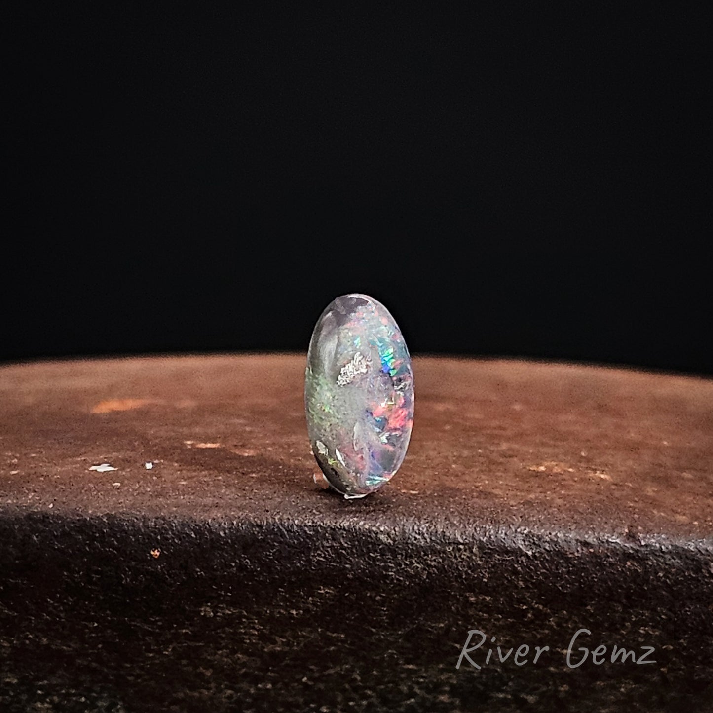 Unset multi-coloured black opal stone on a dark surface with 'River Gemz' branding.