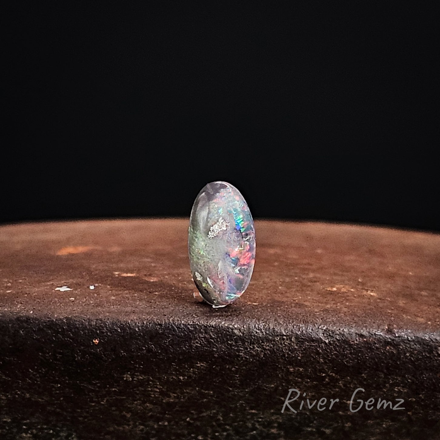 Unset multi-coloured black opal stone on a dark surface with 'River Gemz' branding.