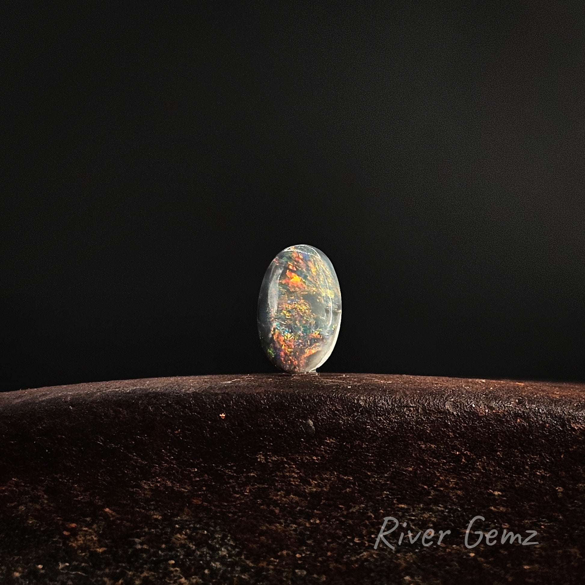 Rare fire colours exhibited in the black opal standing upright on a brown base against a black background.