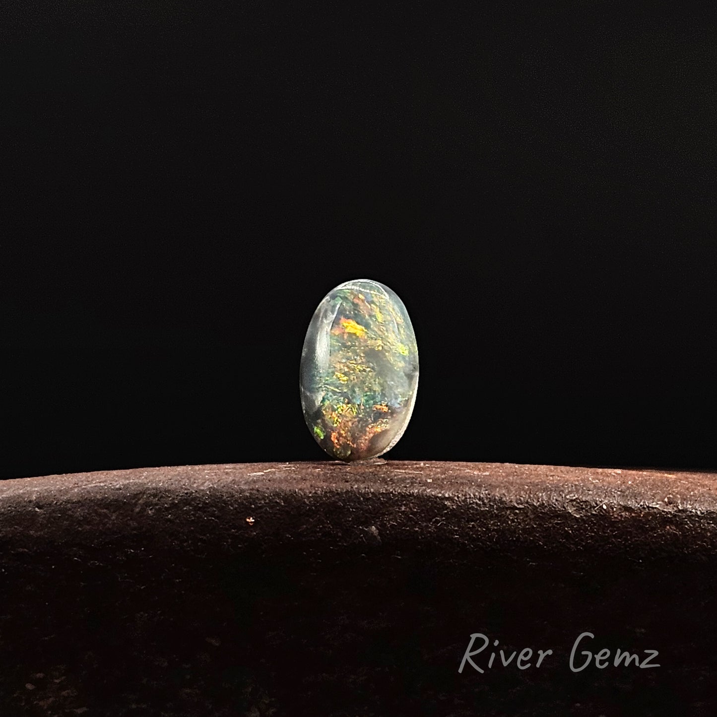 Multi-coloured oval shaped black opal standing against a black background and 'River Gemz' watermark in the bottom right corner.