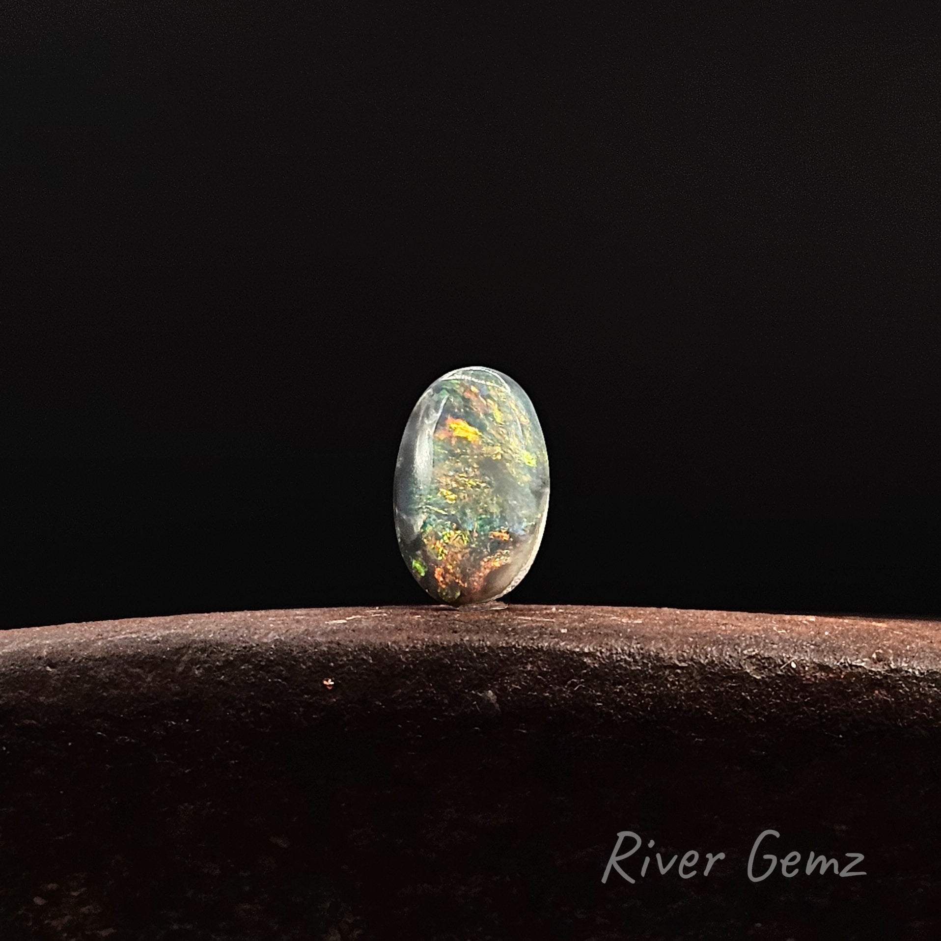 Multi-coloured oval shaped black opal standing against a black background and 'River Gemz' watermark in the bottom right corner.