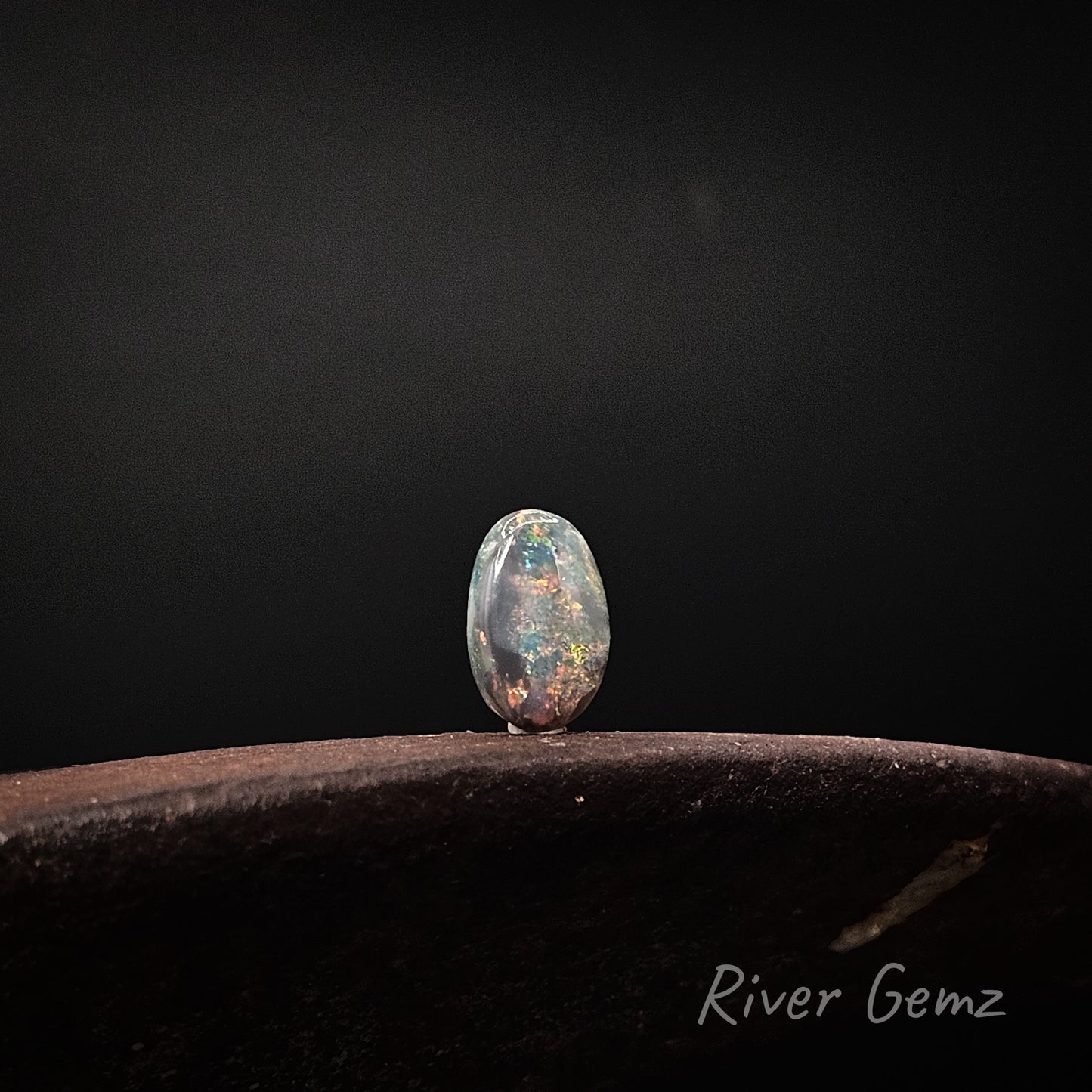 A single oval cabochon gemstone with a vibrant multicolor play-of-colour, standing on a dark textured surface against a dark gray background. The brand name 'River Gemz' is visible in the bottom right corner.